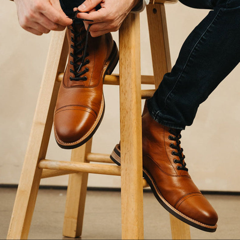 Person sitting on a wooden stool wearing tan leather cap toe lace up boots and a beige jacket.