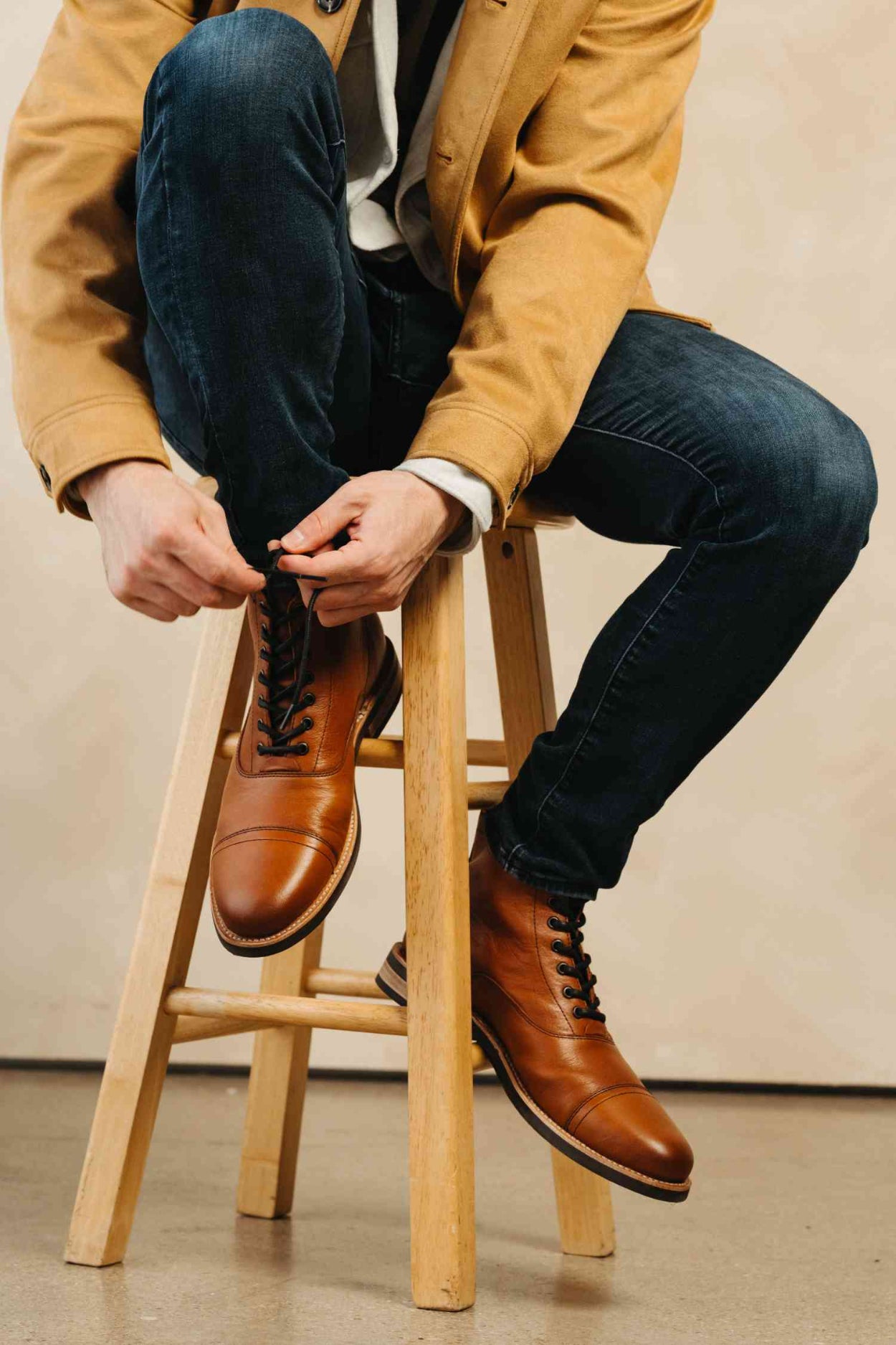 Person sitting on a stool wearing brown leather boots and a mustard jacket.